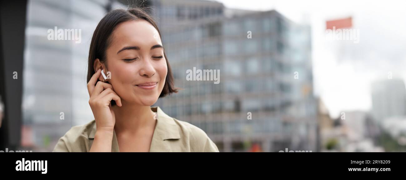 Prise de vue verticale d'une femme asiatique souriante dans un casque sans fil, aime écouter de la musique dans un casque, tient le téléphone portable Banque D'Images