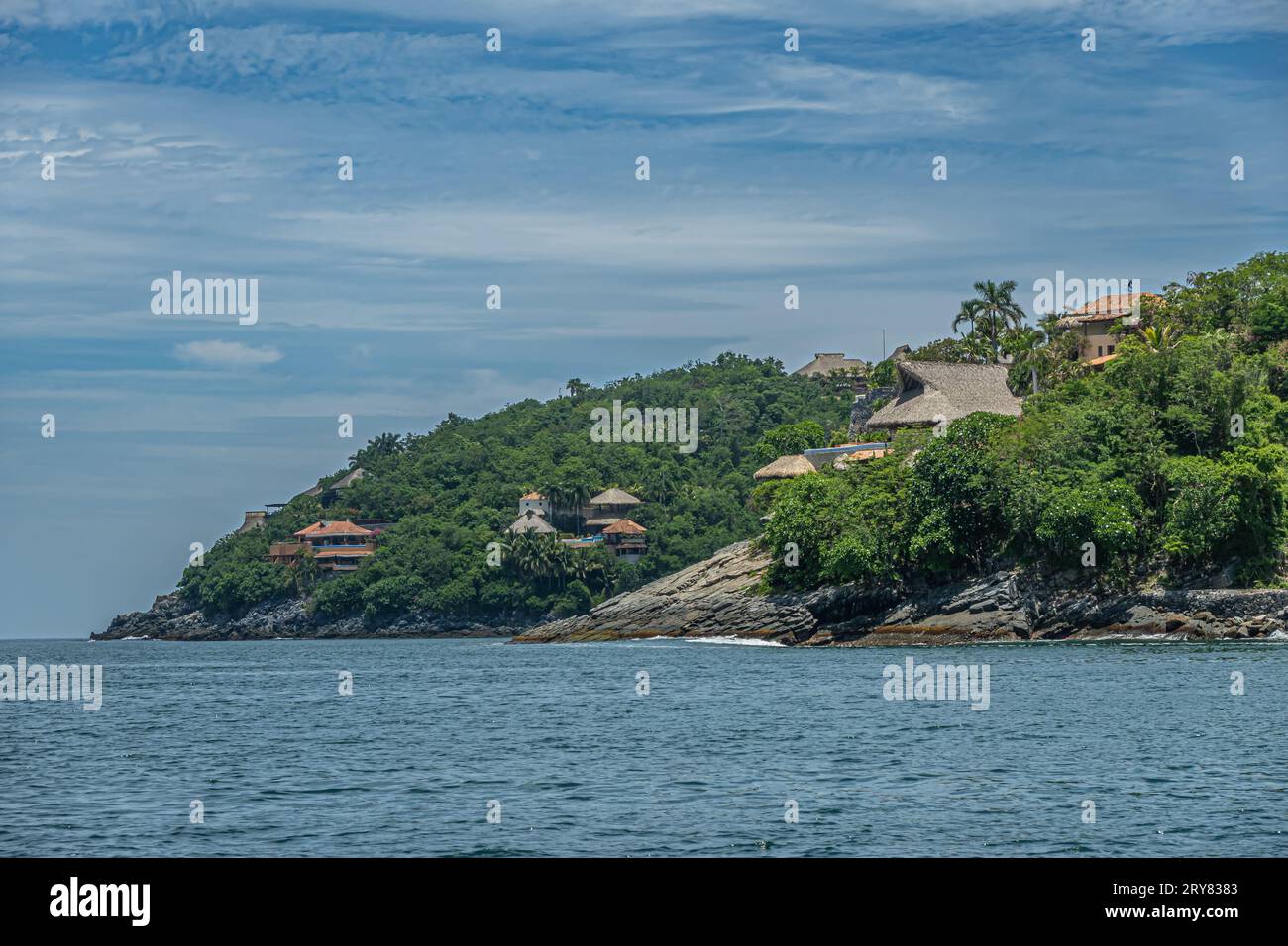 Zihuatanejo, Mexique - 18 juillet 2023 : demeures sur les falaises vertes boisées du rivage de l'océan montrant leurs toits rouges sous le paysage nuageux bleu Banque D'Images