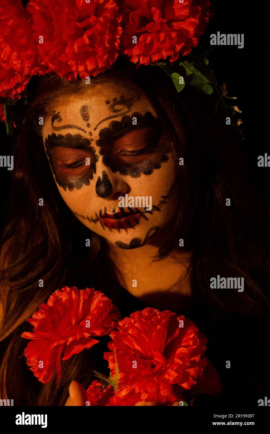 femme habillée en catrina sur un fond sombre regardant un bouquet de fleurs rouges qu'elle tient dans ses mains Banque D'Images