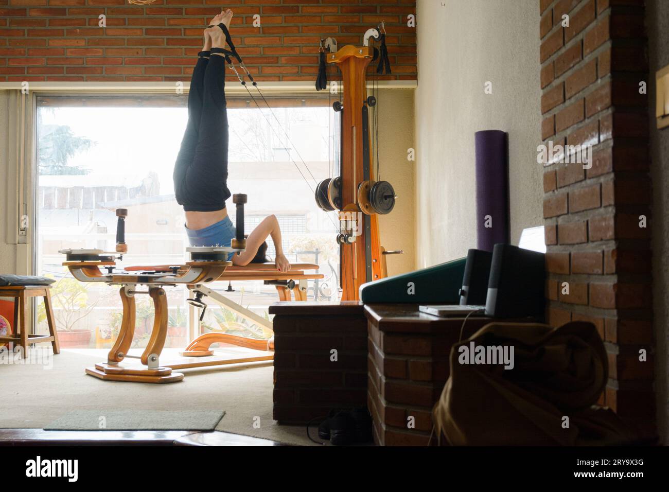Femme caucasienne avec des vêtements de sport bleus, dans le salon de son appartement formant son énergie et son équilibre avec la machine d'exercice, en utilisant des poulies avec fenêtre Banque D'Images