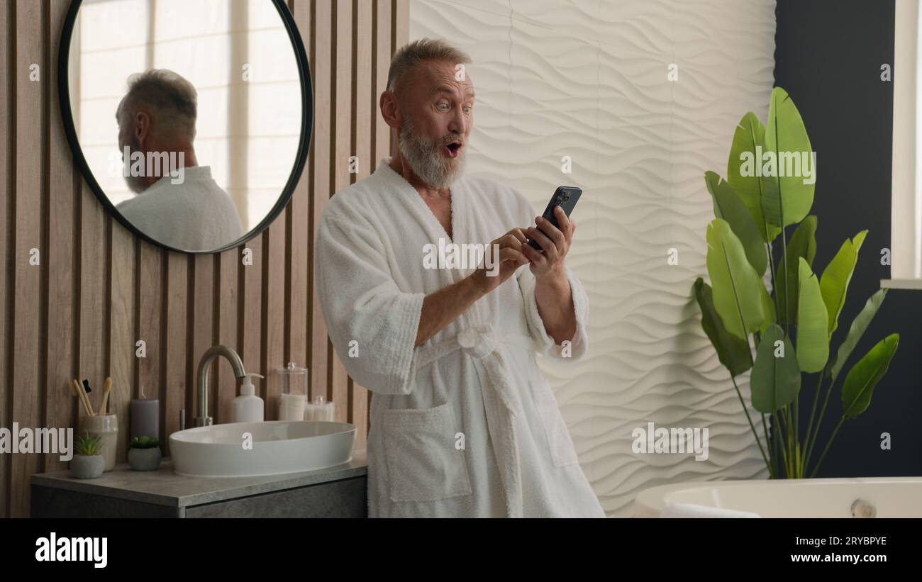 Caucasien heureux vieux retraité homme dans la salle de bains âgé senior mature mâle dans le bain hygiène du matin regardant téléphone portable lisant de bonnes nouvelles en ligne Banque D'Images