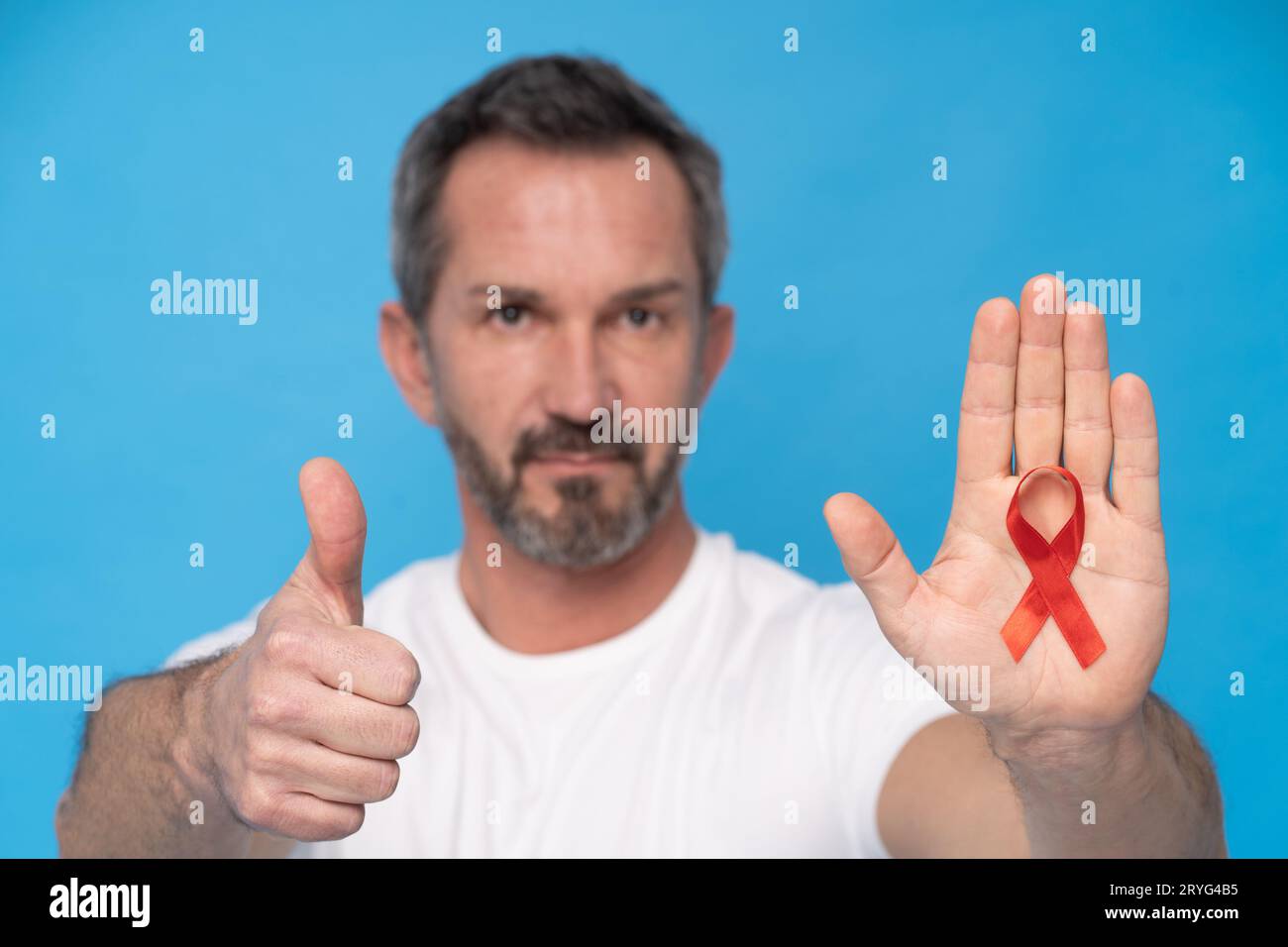 Pouce vers le haut geste homme mature avec ruban rouge symbole de sensibilisation SIDA sur une paume portant un t-shirt blanc isolé sur un dos bleu Banque D'Images