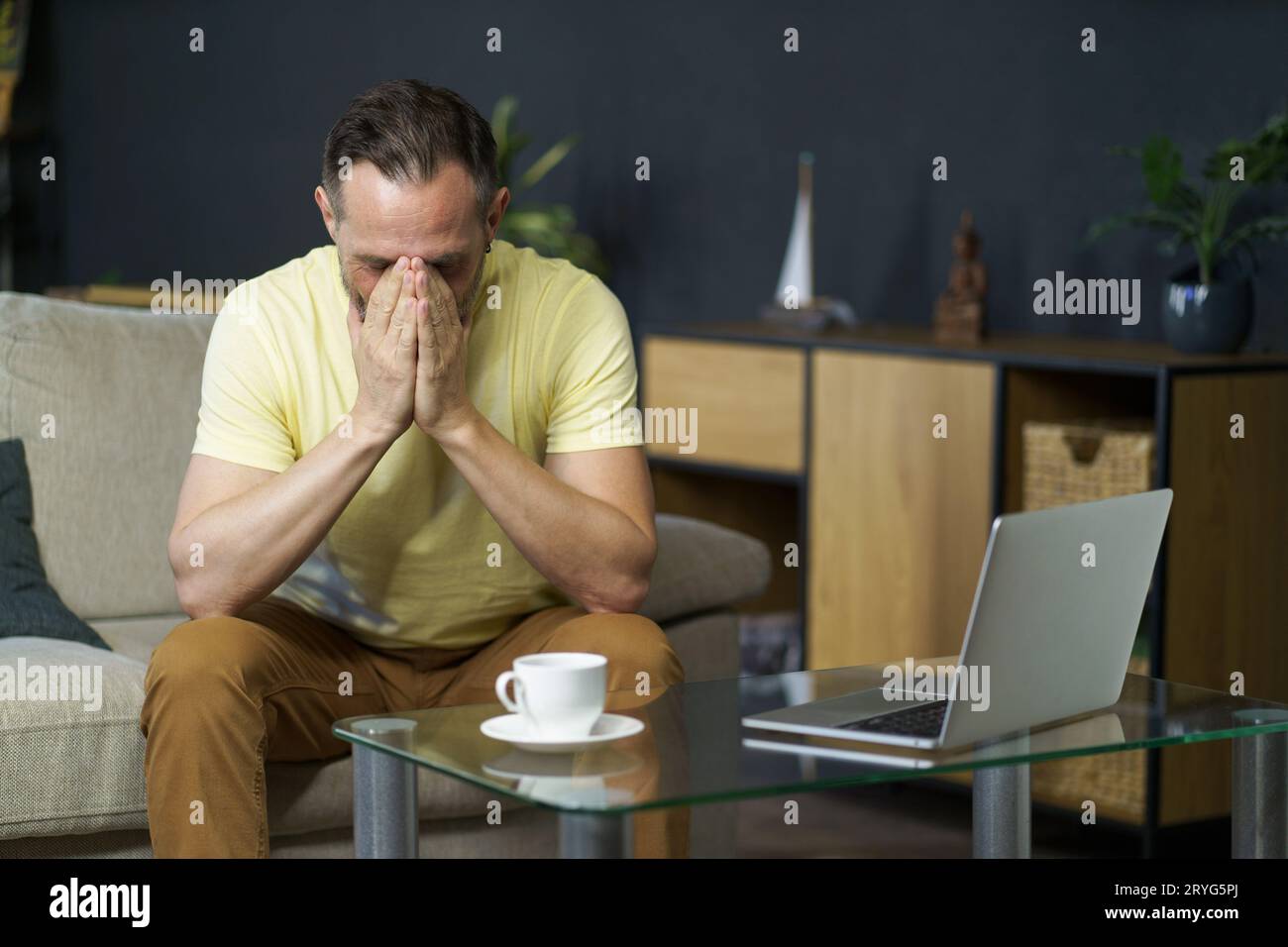 Crise d'âge moyen, stress émotionnel homme mature assis sur le canapé avec les mains couvertes son visage à côté de la table basse avec ordinateur portable et tasse Banque D'Images