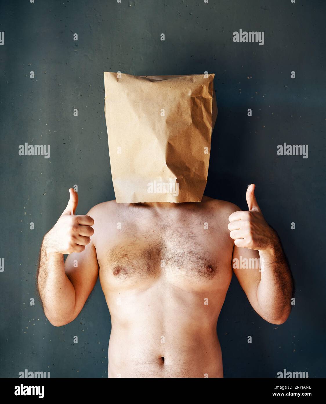 Homme torse nu avec un sac en papier sur la tête montrant les pouces vers le haut sur fond gris Banque D'Images