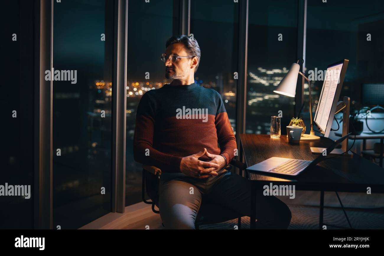 Un homme d'affaires caucasien pensif travaillant tard dans le bureau. Il est assis à son bureau, absorbé par son ordinateur portable, regardant loin dans la nuit, pensant à Banque D'Images