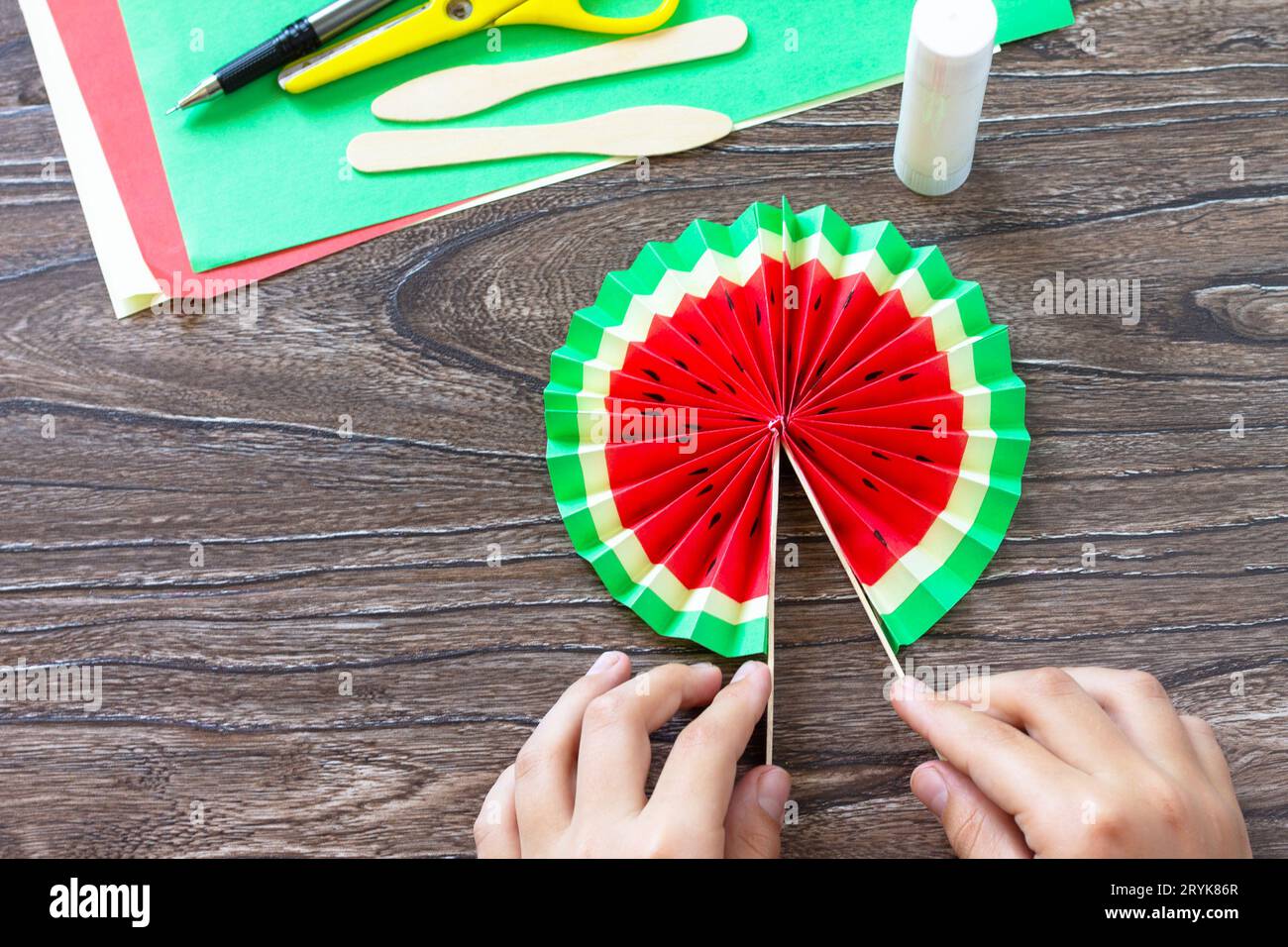 Dans les mains d'un enfant papier ventilateur pastèque sur une table en bois. Projet d'art pour enfants, fait à la main, artisanat pour enfants. Banque D'Images