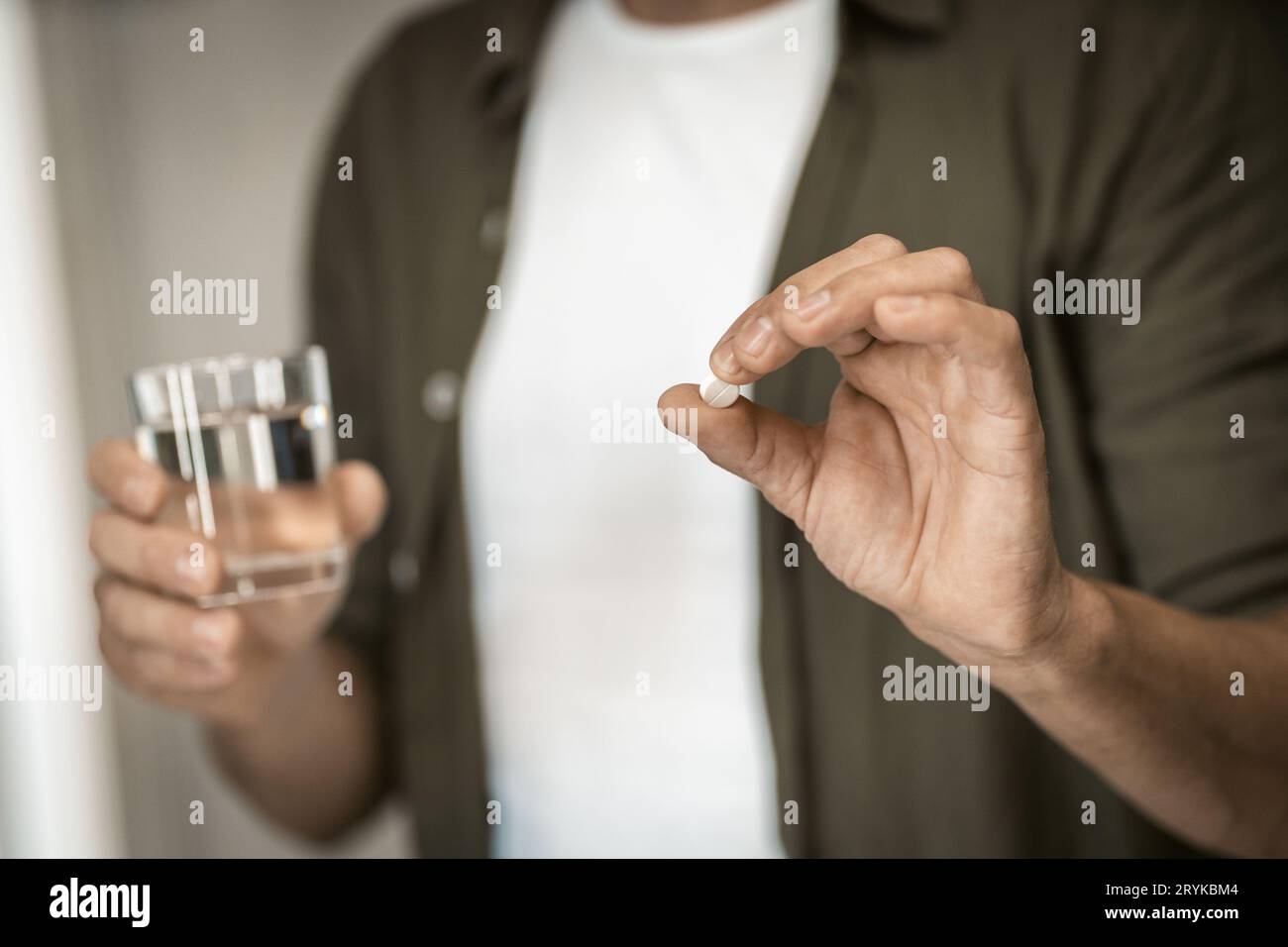 Gros plan d'un homme tenant une pilule dans une main et un verre d'eau dans l'autre, dans le confort de sa maison. La mise au point douce sélective attire l'attention Banque D'Images