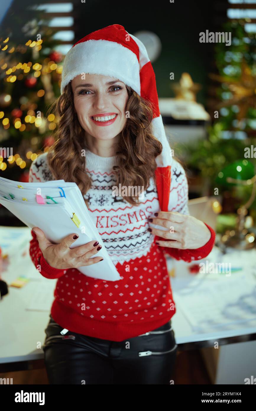 Noël. Portrait de souriant élégante femme propriétaire de petite entreprise de 40 ans dans le chapeau de santa et pull de Noël rouge avec des documents en gr moderne Banque D'Images