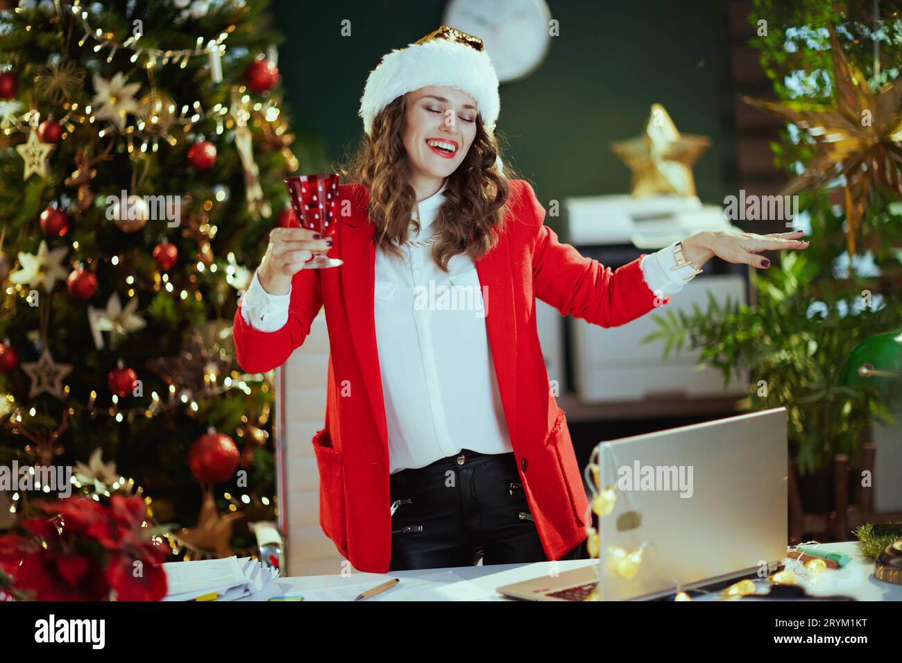 Noël. Heureuse élégante femme d'affaires d'âge moyen dans le chapeau de santa et veste rouge avec le verre de vin dansant dans le bureau vert moderne avec l'arbre de Noël Banque D'Images