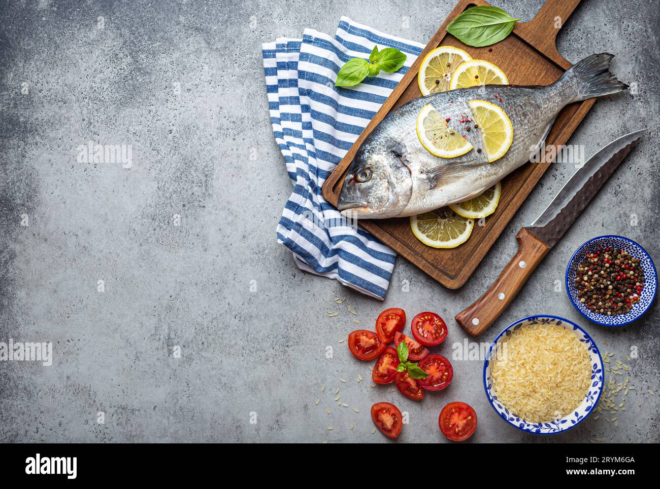 Poisson cru dorado avec des ingrédients citron, basilic frais, tomates cerises coupées, riz cru sur planche à découper en bois avec couteau sur RU Banque D'Images