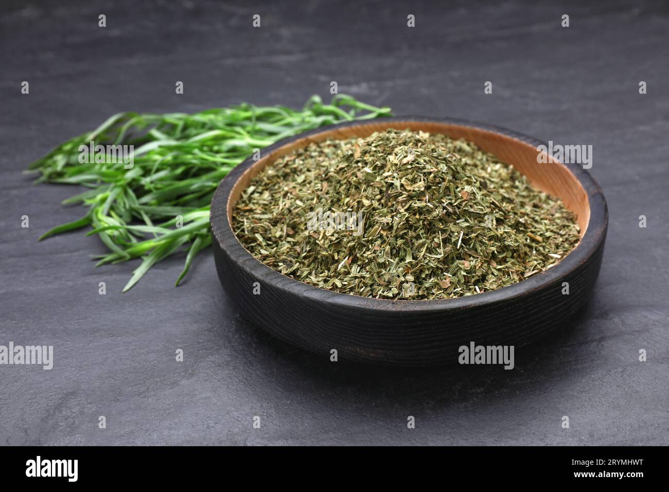 Sécher l'estragon dans un bol et les feuilles vertes sur la table texturée noire, gros plan Banque D'Images