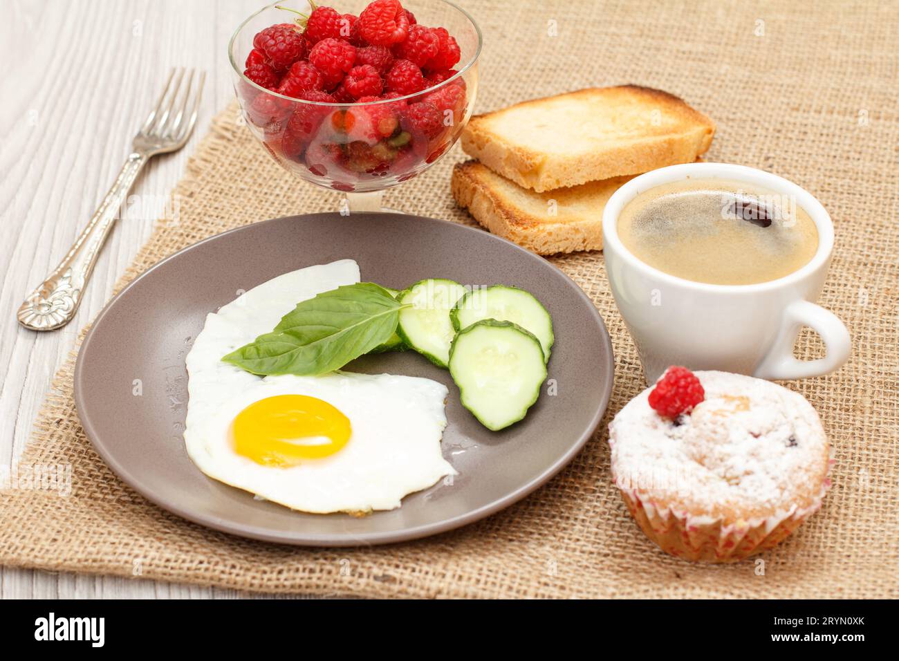 Assiette avec œuf au plat, concombre coupé et feuille de basilic, muffin, tasse de café noir, toasts Banque D'Images