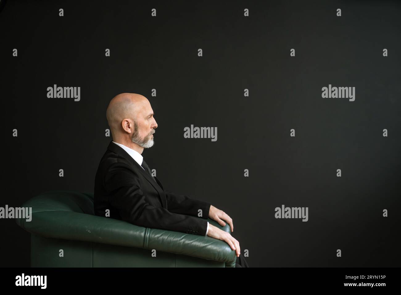 Homme mature dans un costume, assis dans une chaise en cuir confortable. La toile de fond d'un mur noir ajoute une touche d'élégance et de sophisti Banque D'Images