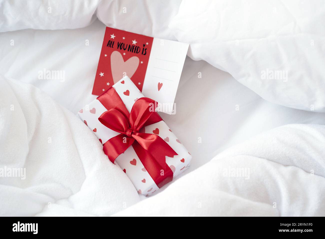 Boîte cadeau dans un emballage en papier avec des coeurs et un noeud de ruban rouge et une carte de Saint-Valentin sur un lit tôt le matin. Surprise pour Banque D'Images