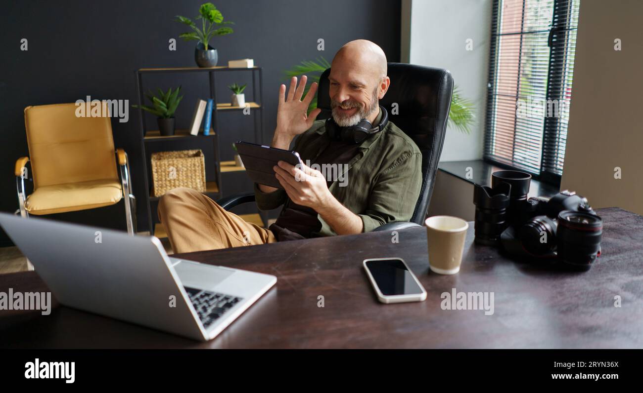 Le photographe a un appel en ligne avec le client. Il détient Tablet PC et interlocuteur accueillant. Bureau à l'avant équipé avec ordinateur portable, mob Banque D'Images