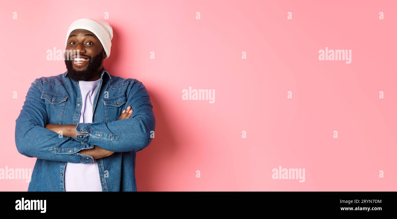 Heureux Noir homme à la barbe, portant un bonnet et une chemise en denim, l'air intrigué et amusé à la caméra, souriant avec les bras croisés Banque D'Images