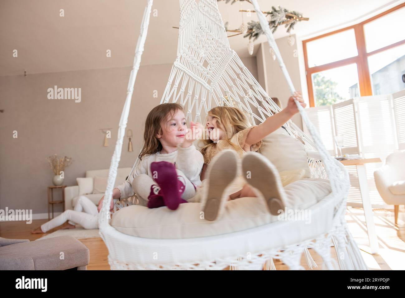 Deux petites filles montent sur une balançoire macramé à la maison près du sapin de Noël. Les sœurs s'amusent, se trompent en vacances. Amies drôles ayant un hap Banque D'Images