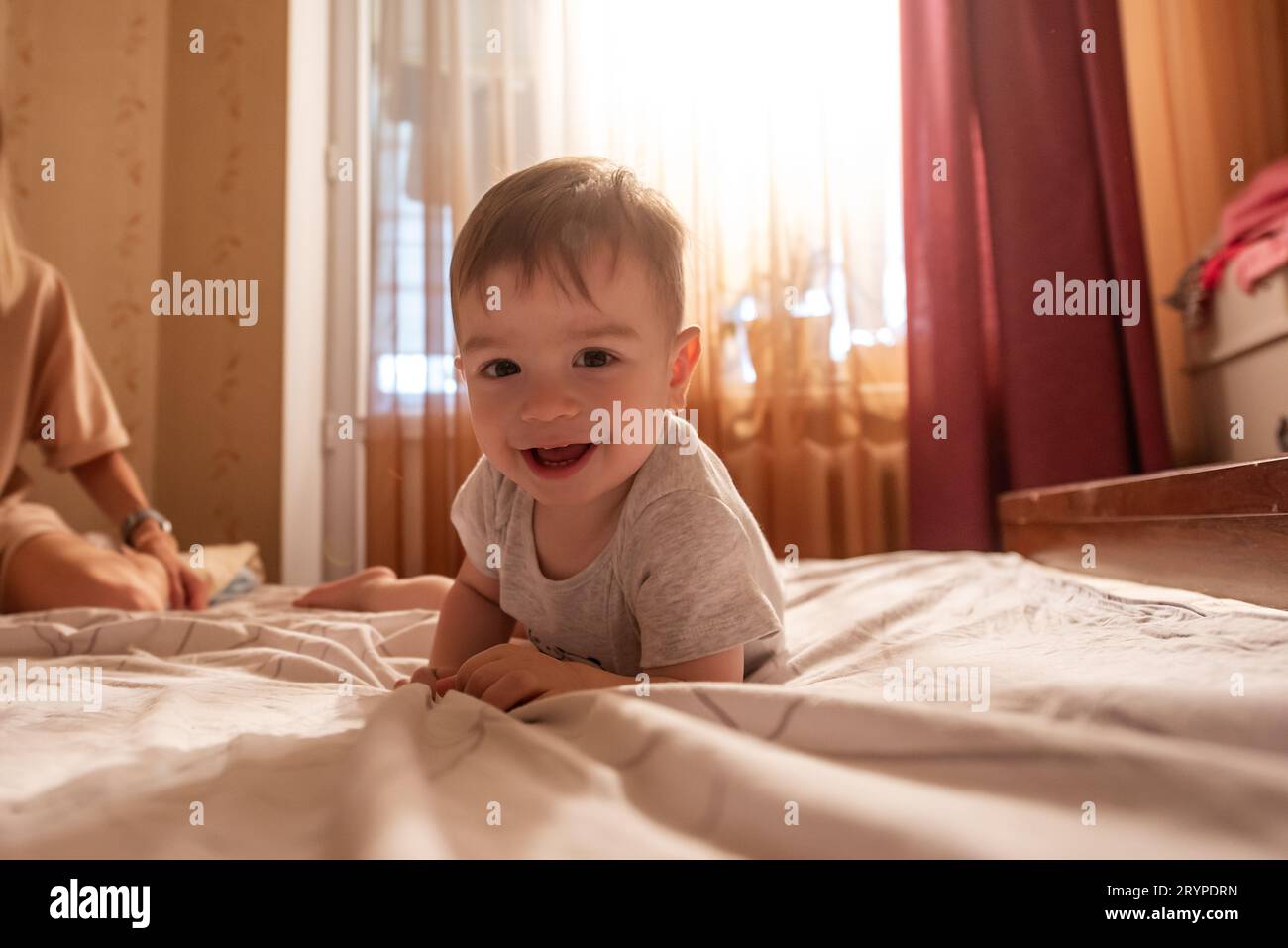 Bébé d'un an allongé sur le ventre sur le lit le matin au soleil. L'enfant est drôle, souriant, jouant de bonne humeur. Soins, meubles pour Banque D'Images