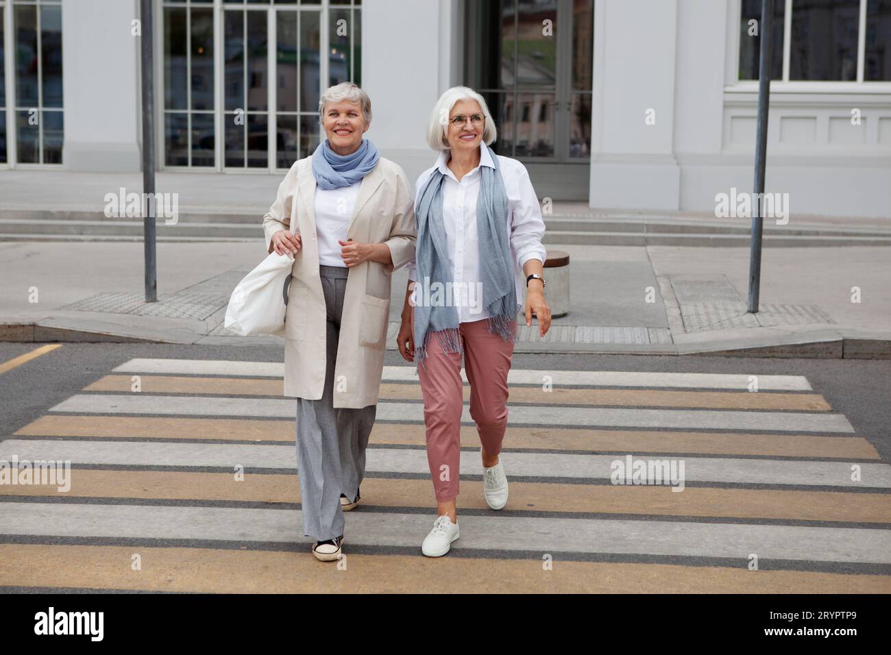 Deux femmes âgées aux cheveux gris dans des vêtements élégants, pantalons, manteaux, baskets marchant, traversant la route en ville et sourires. Femme âgée à la mode Banque D'Images