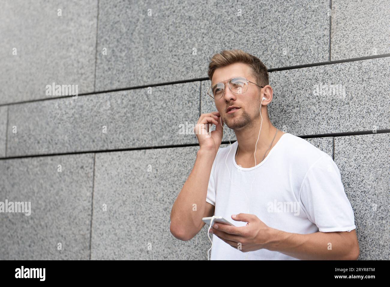 Mec gai habillé en t-shirt blanc dans la rue, écoutant de la musique avec des écouteurs, tenant le téléphone portable. Banque D'Images