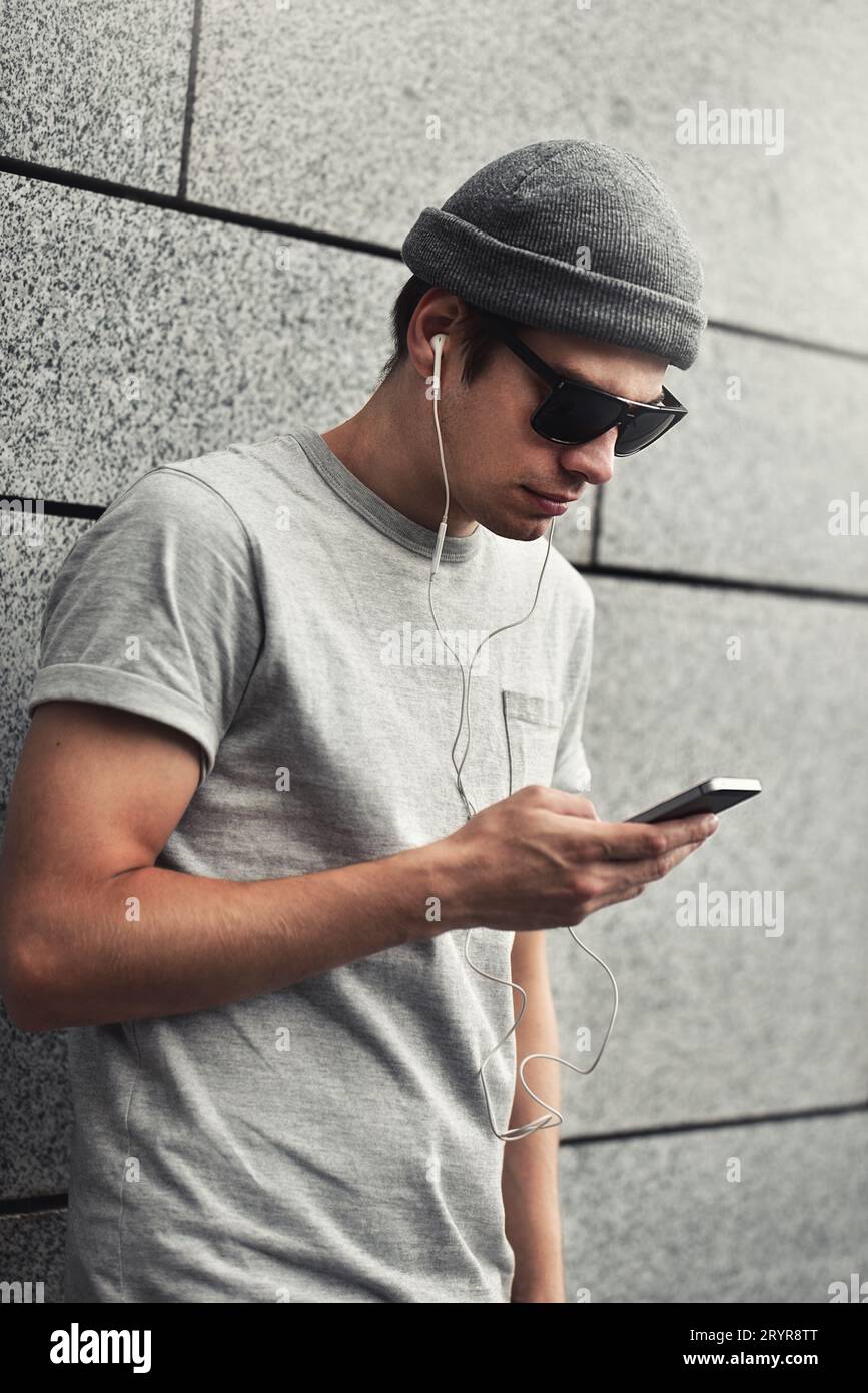 Mec gai habillé en t-shirt gris, lunettes de soleil et chapeau dans la rue, écoutant de la musique avec des écouteurs, tenant le téléphone portable. Banque D'Images