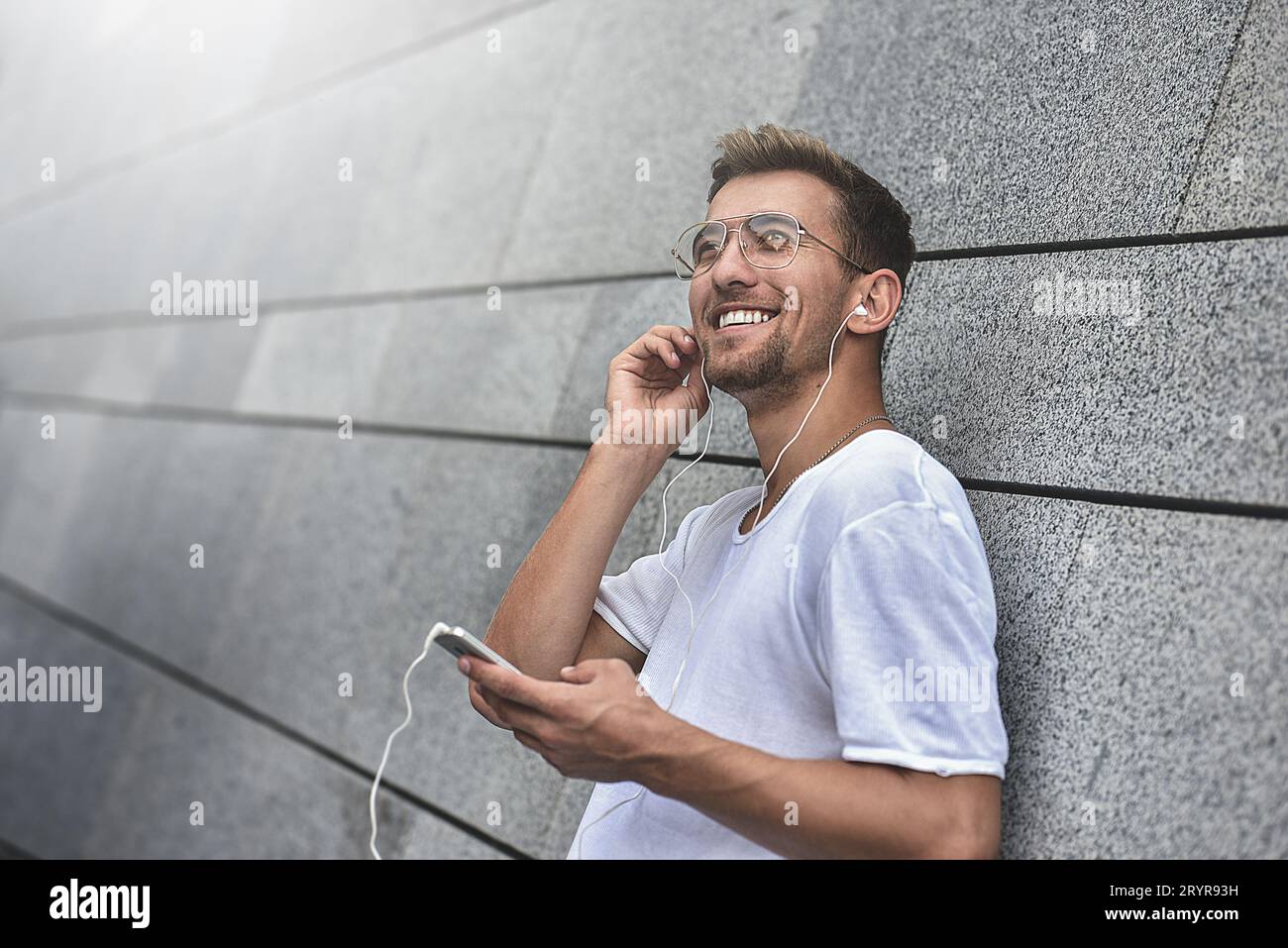 Mec gai habillé en t-shirt blanc dans la rue, écoutant de la musique avec des écouteurs, tenant le téléphone portable. Banque D'Images