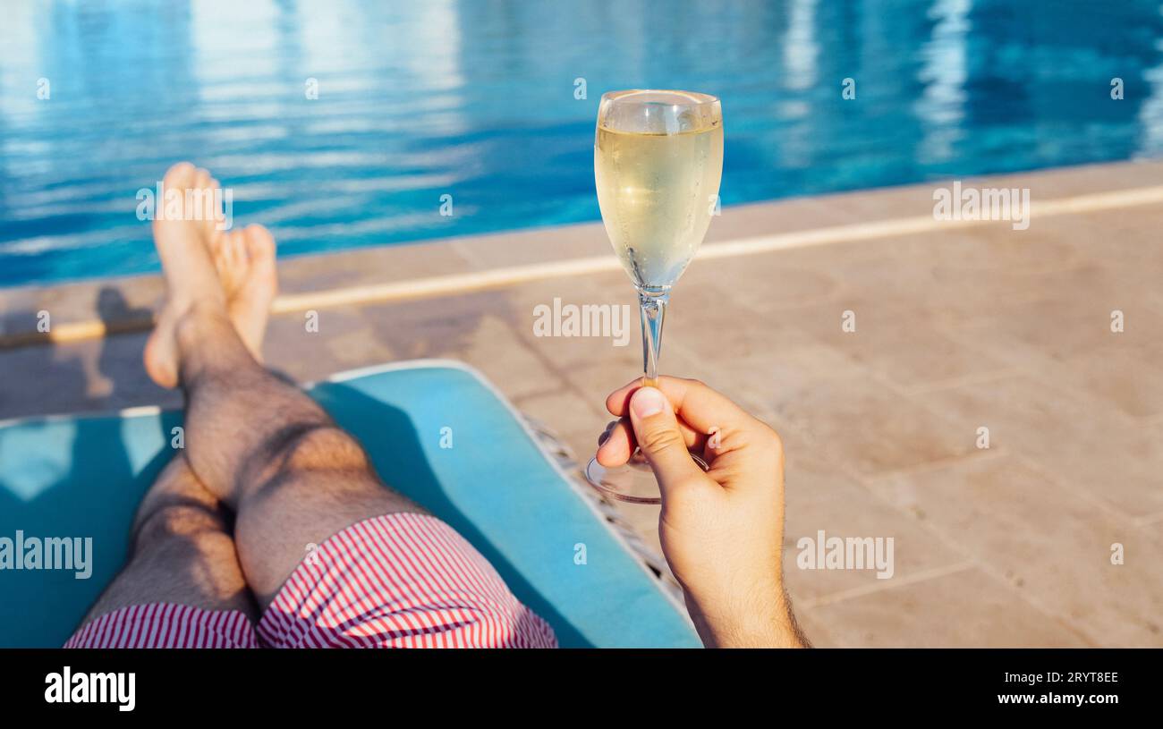 Jeune homme en short se trouve sur une chaise longue et tient un élégant verre de champagne frais avec une longue tige. Banque D'Images
