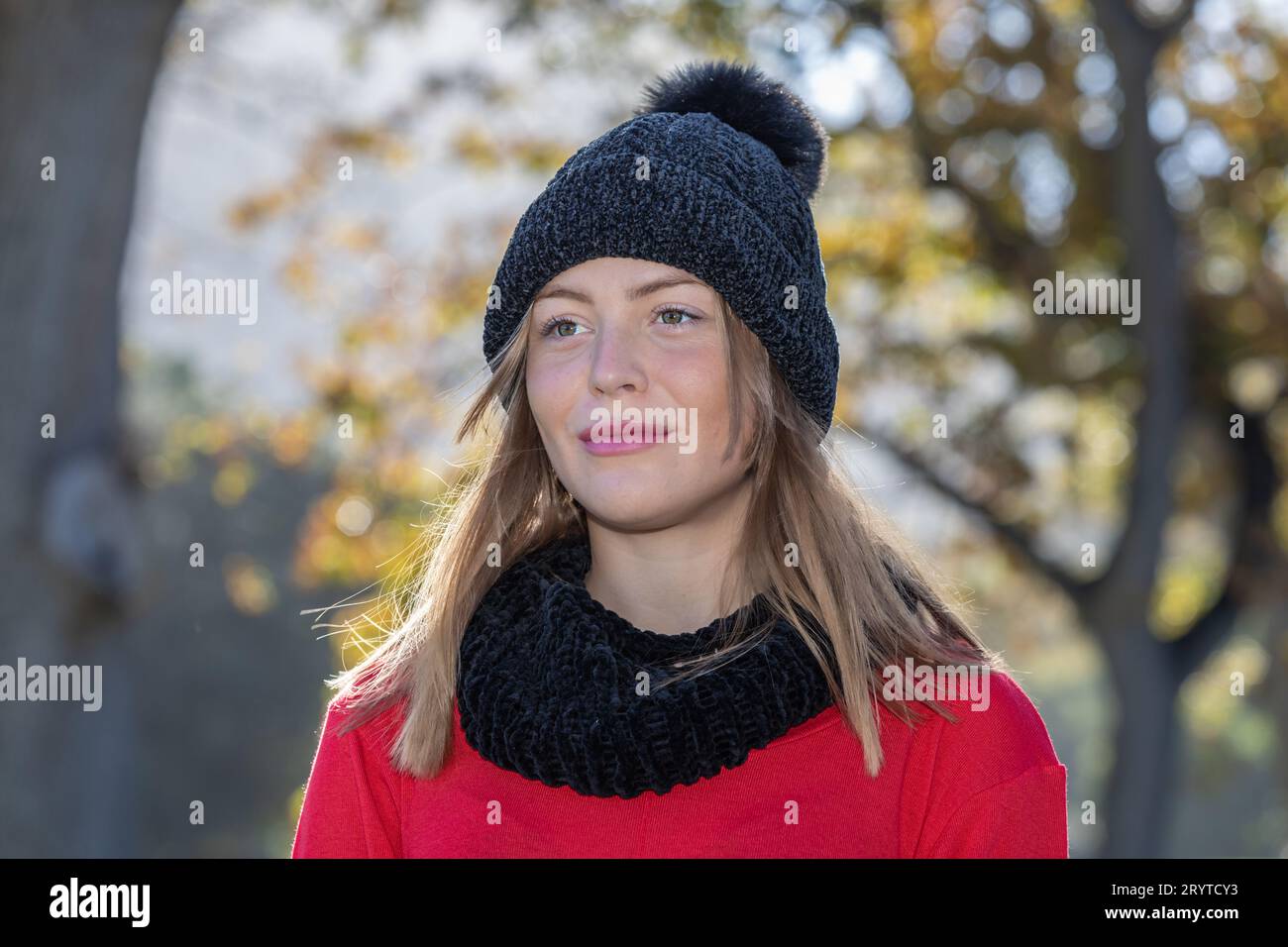 Dans une photo de la tête et des épaules, une superbe jeune femme blonde, portant un chapeau de laine noir et un pull rouge vif, rayonne sous les arbres d'automne Banque D'Images