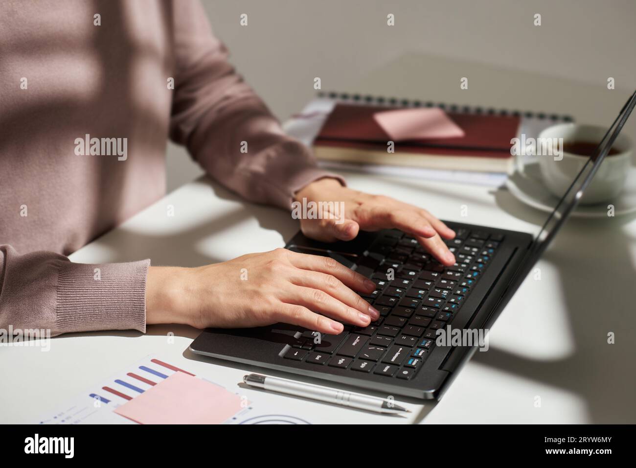 Mains de la jeune femme économiste en pull beige tapant sur le clavier d'ordinateur portable tout en étant assis par le bureau avec des documents financiers et stylo Banque D'Images