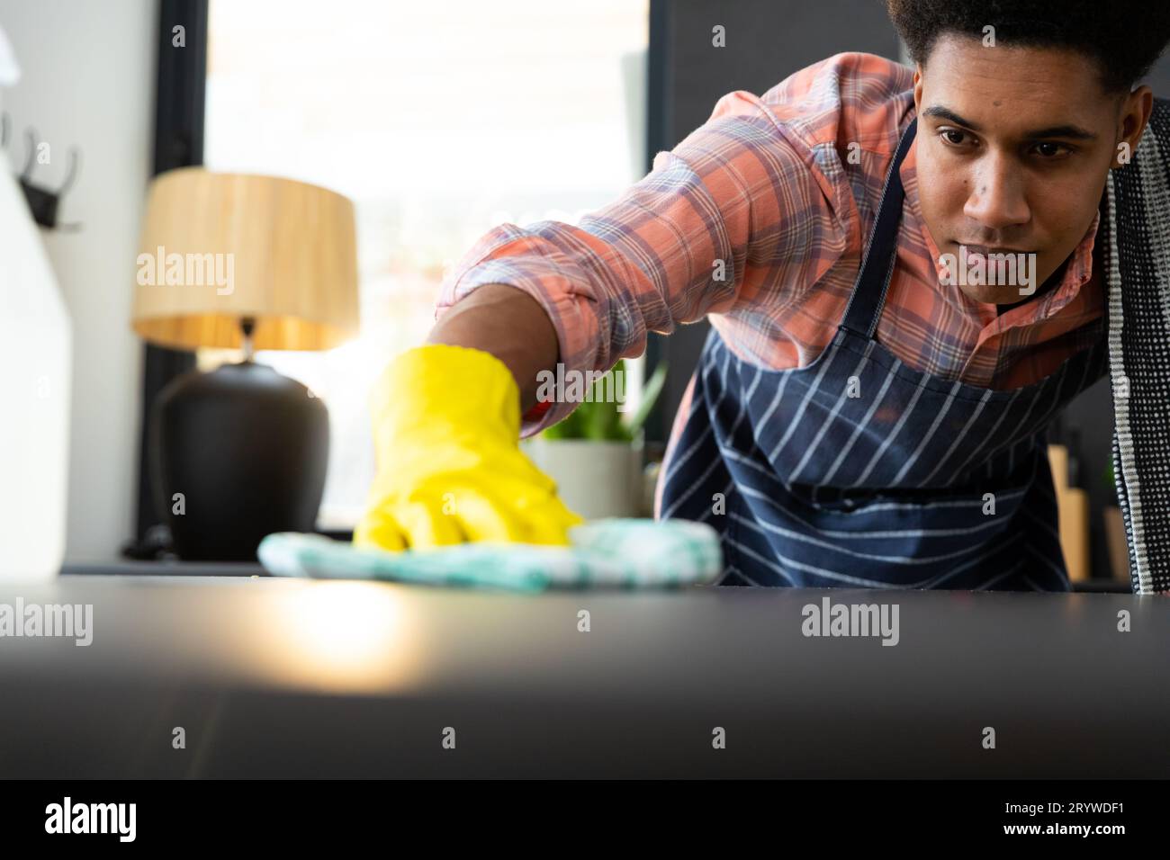 Homme biracial concentré portant un tablier et des gants en caoutchouc nettoyant le comptoir dans la cuisine ensoleillée Banque D'Images