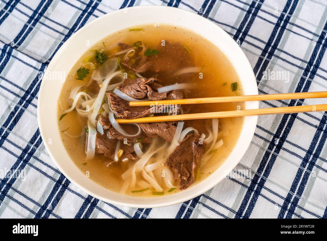 Soupe épicée vietnamienne traditionnelle Pho Bo avec morceau de bœuf, nouilles de riz et herbes dans une assiette avec baguette sur une nappe à carreaux, gros plan. Banque D'Images