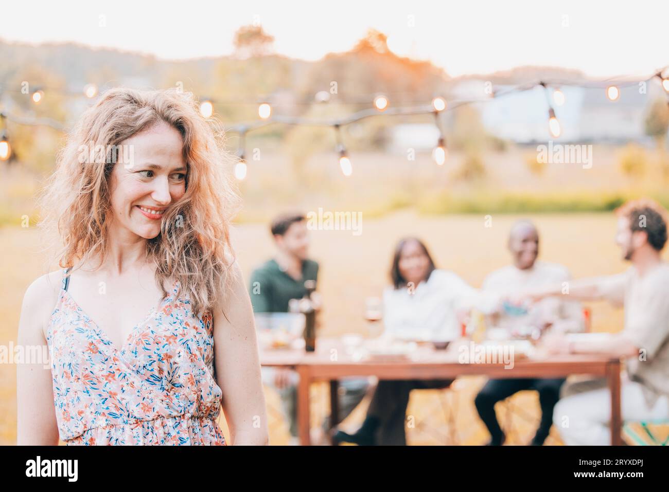 Belle jeune femme avec les cheveux blonds bouclés dans une robe d'été avec un groupe d'amis en arrière-plan ayant un jardin extérieur Banque D'Images