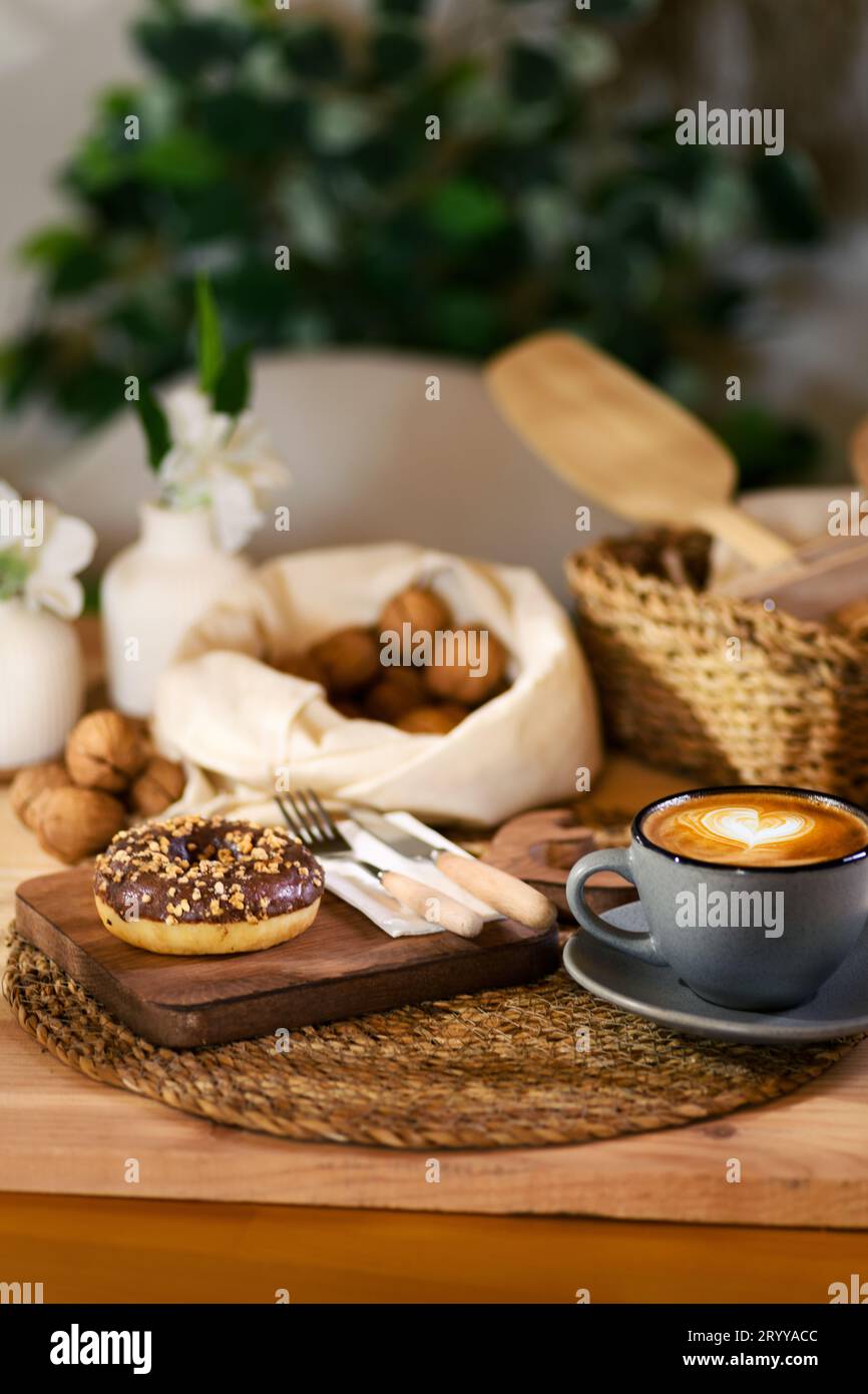 Un beignet sur une planche de bois, à côté d'un latte dans une tasse grise. Serviette blanche avec couverts en bois et noix décortiquées en tissu blanc à l'arrière-plan. Banque D'Images