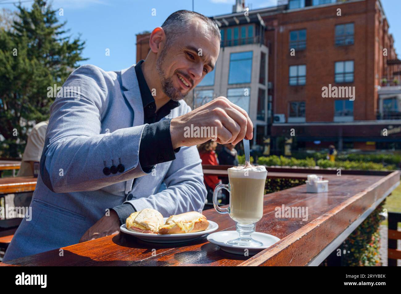 Homme adulte français caucasien avec la barbe cadre d'affaires prenant le petit déjeuner devant le restaurant assis au comptoir, mélangeant le café pour le boire et manger Banque D'Images
