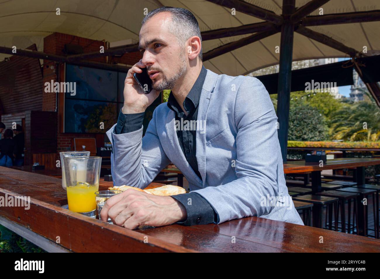 Homme d'affaires caucasien adulte avec barbe et cheveux courts, concentré à parler au téléphone tout en prenant le petit déjeuner assis à l'extérieur du restaurant pendant su Banque D'Images