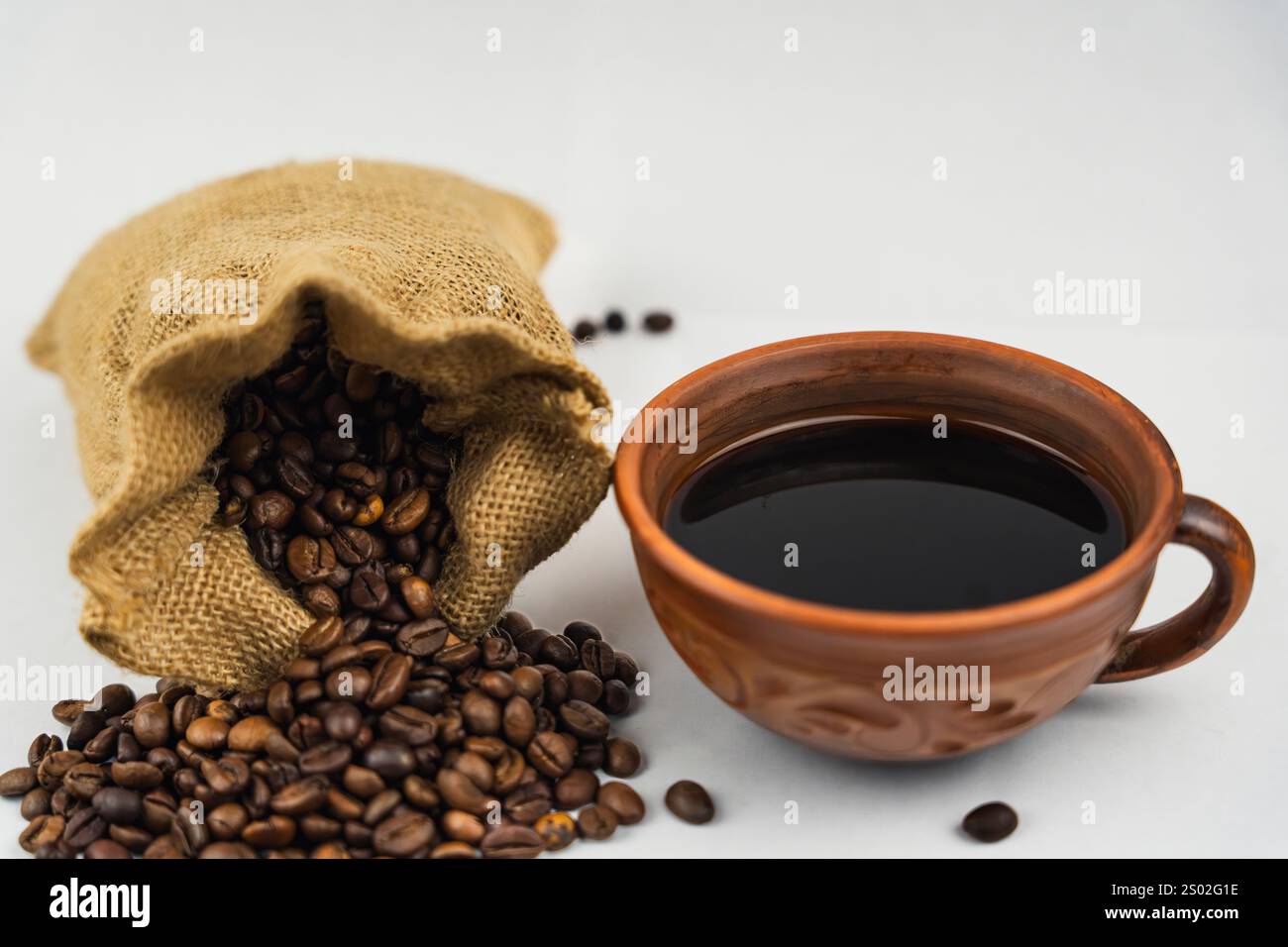Grains de café renversant du sac de toile de jute à côté d'une tasse d'argile remplie de café noir sur fond blanc, style rustique et naturel. Banque D'Images
