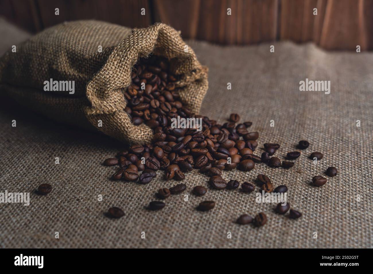Un sac en toile de jute renversant des grains de café torréfiés sur une surface texturée, capturant un thème de café naturel et rustique. Banque D'Images