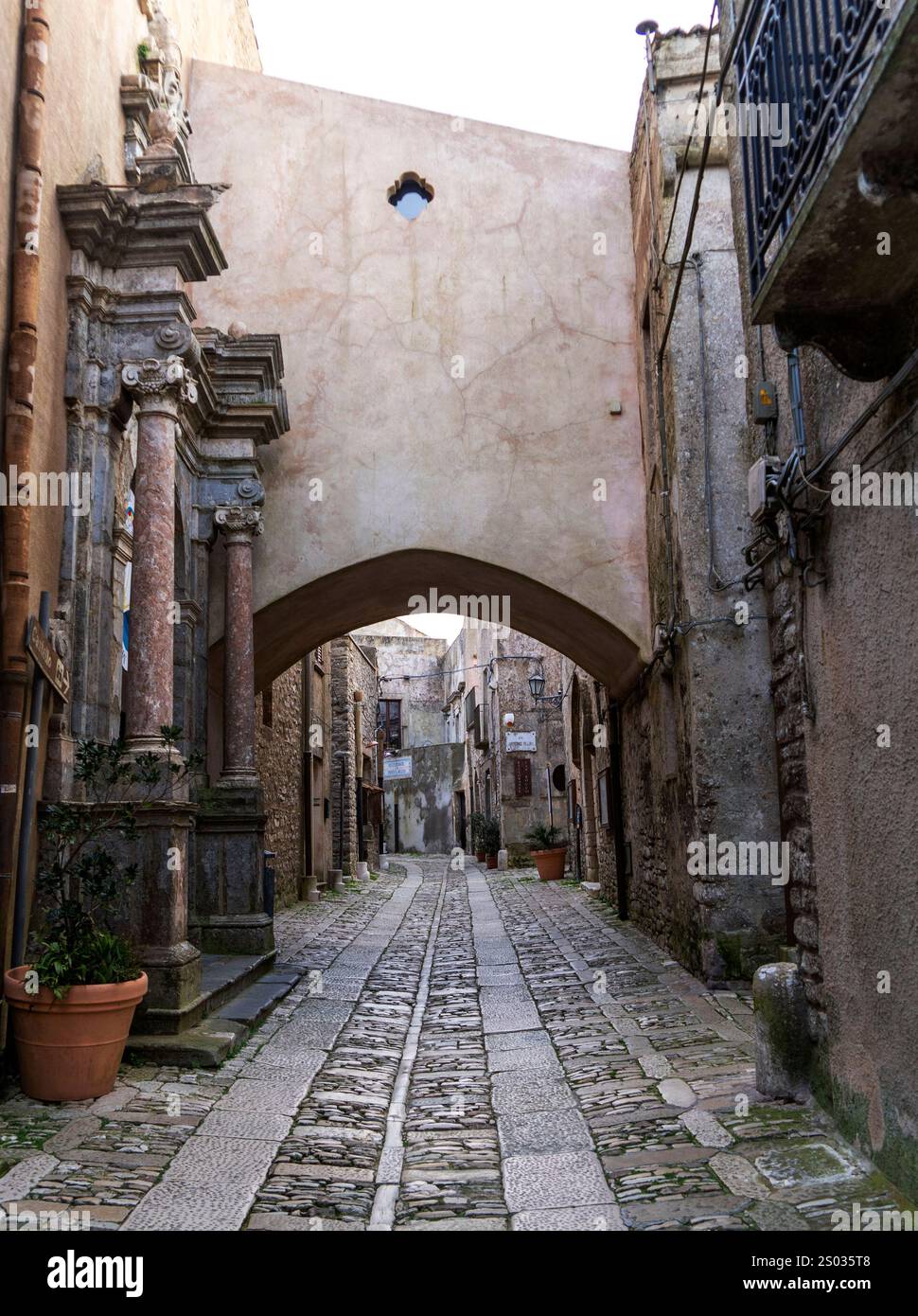 Une étroite rue pavée dans une vieille ville, avec une arche et une architecture historique. Des plantes en pot bordent la rue, et les bâtiments montrent des signes Banque D'Images
