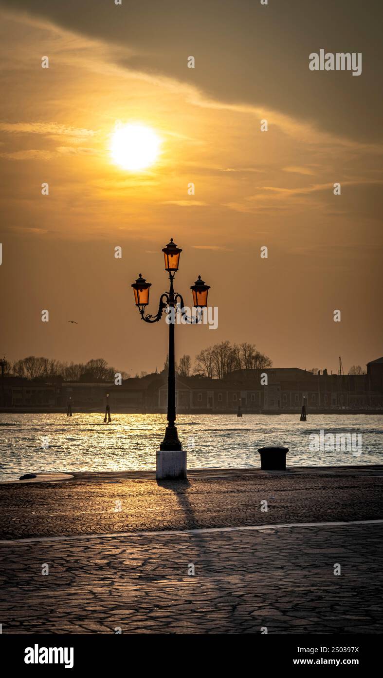 Un coucher de soleil serein sur un plan d'eau, avec un lampadaire vintage au premier plan. Le soleil projette une lueur chaude, éclairant la scène avec un l doux Banque D'Images