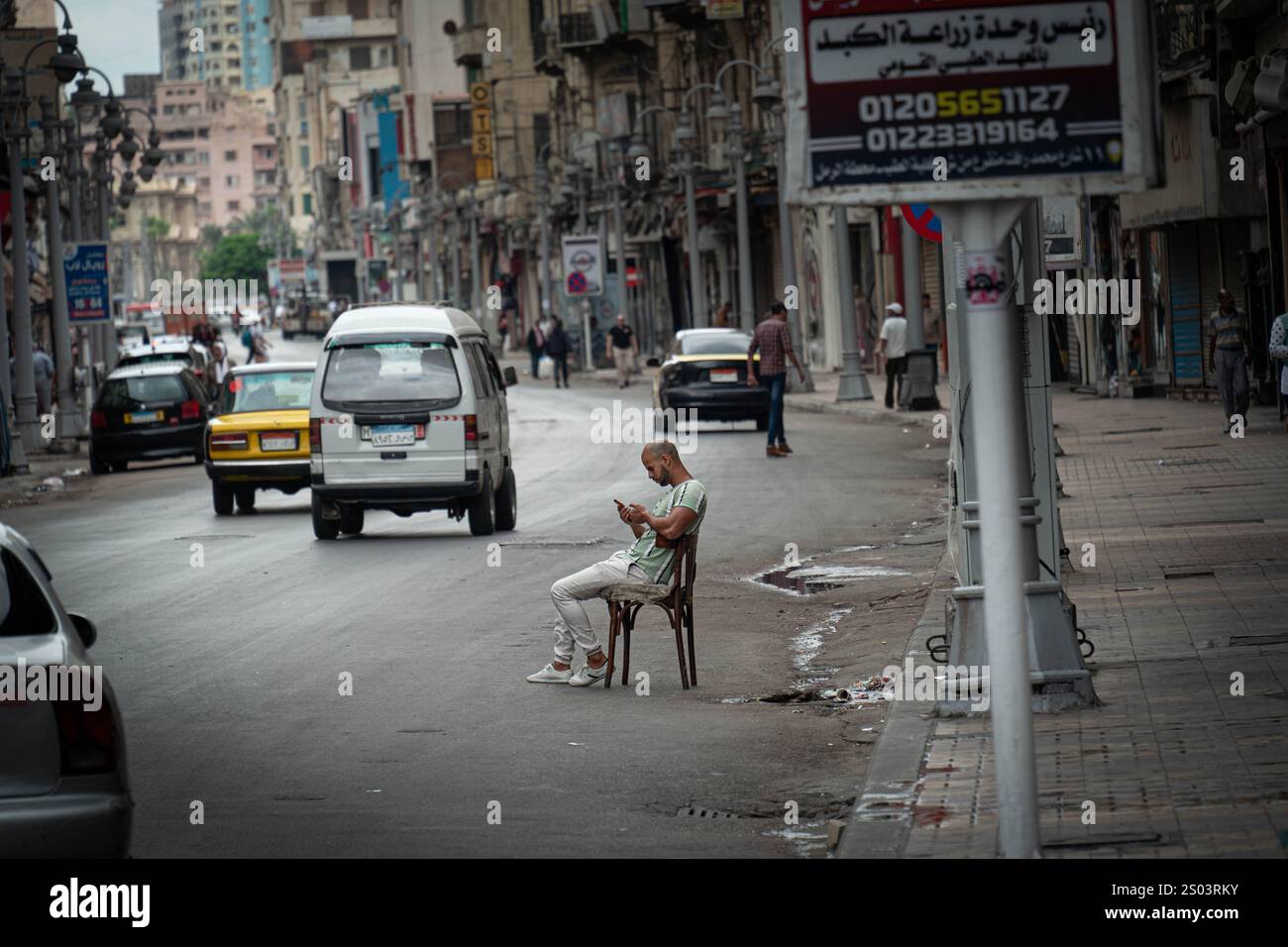 Homme assis sur une chaise au milieu d'une rue calme à Alexandrie, Egypte, immergé dans son téléphone. Trafic environnant et environnement urbain. Banque D'Images