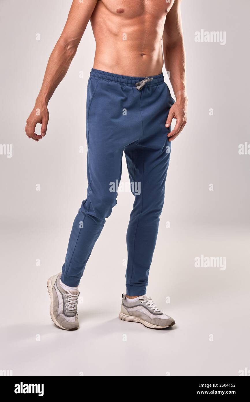 Homme sportif dans des joggers bleus à la mode, démontrant des vêtements décontractés et des vêtements athlétiques, debout sur blanc, concept de mode Banque D'Images