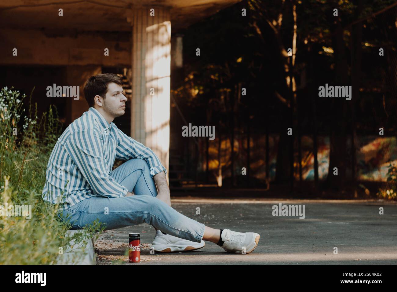 Assis sur un trottoir, un homme en vêtements décontractés de couleur claire regarde au loin avec une expression pensive. Le cadre transmet la tranquillité et le profou Banque D'Images