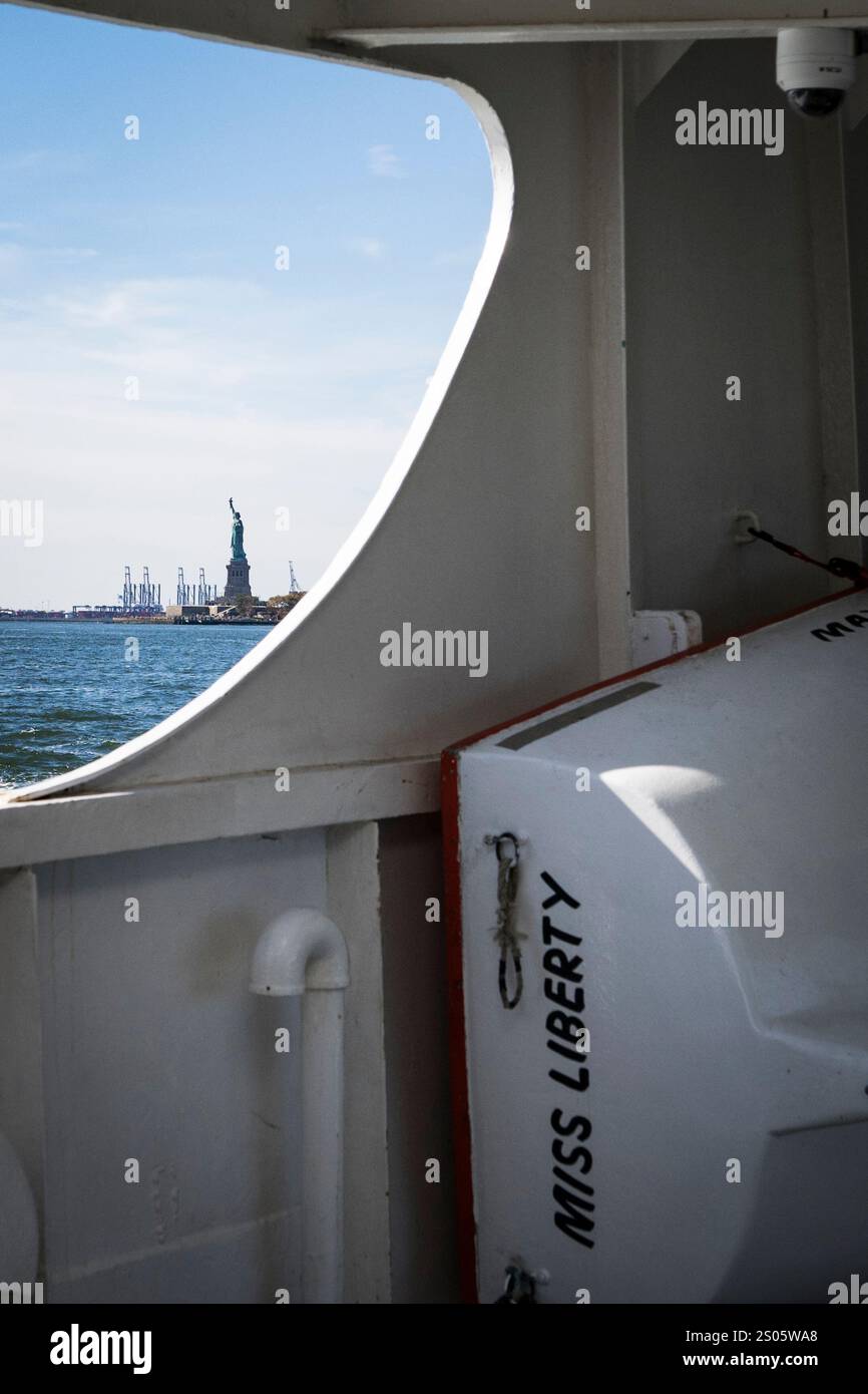 Encadrée par une fenêtre de ferry, Lady Liberty se dresse haut, avec un bateau de sauvetage bien nommé « Miss Liberty » ci-dessous, un hommage parfait à la liberté et à la résilience. Banque D'Images