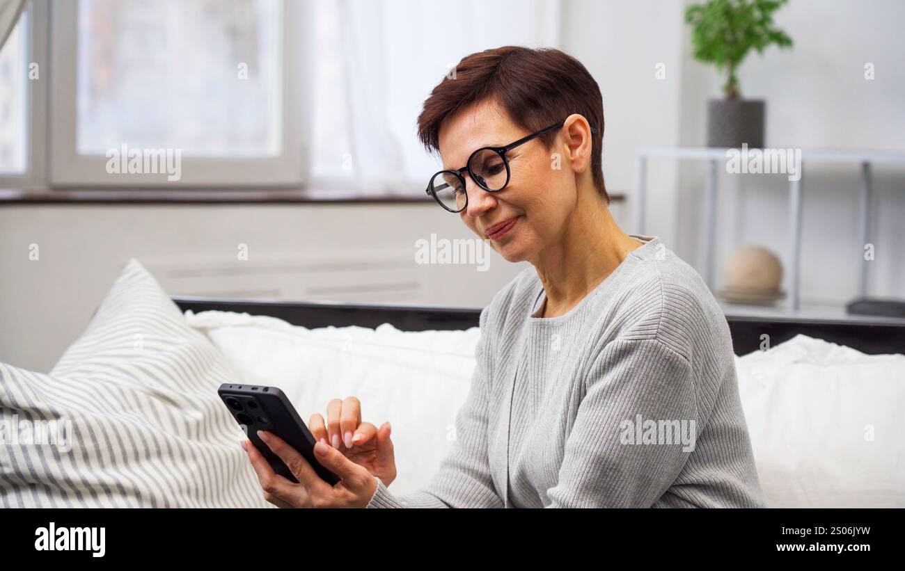 Femme mature avec des lunettes assis sur le canapé à l'aide d'un smartphone pour faire un achat en ligne dans un salon confortable Banque D'Images Femme mature avec des lunettes assis sur le canapé à l'aide d'un smartphone pour faire un achat en ligne dans un salon confortable Banque D'Images