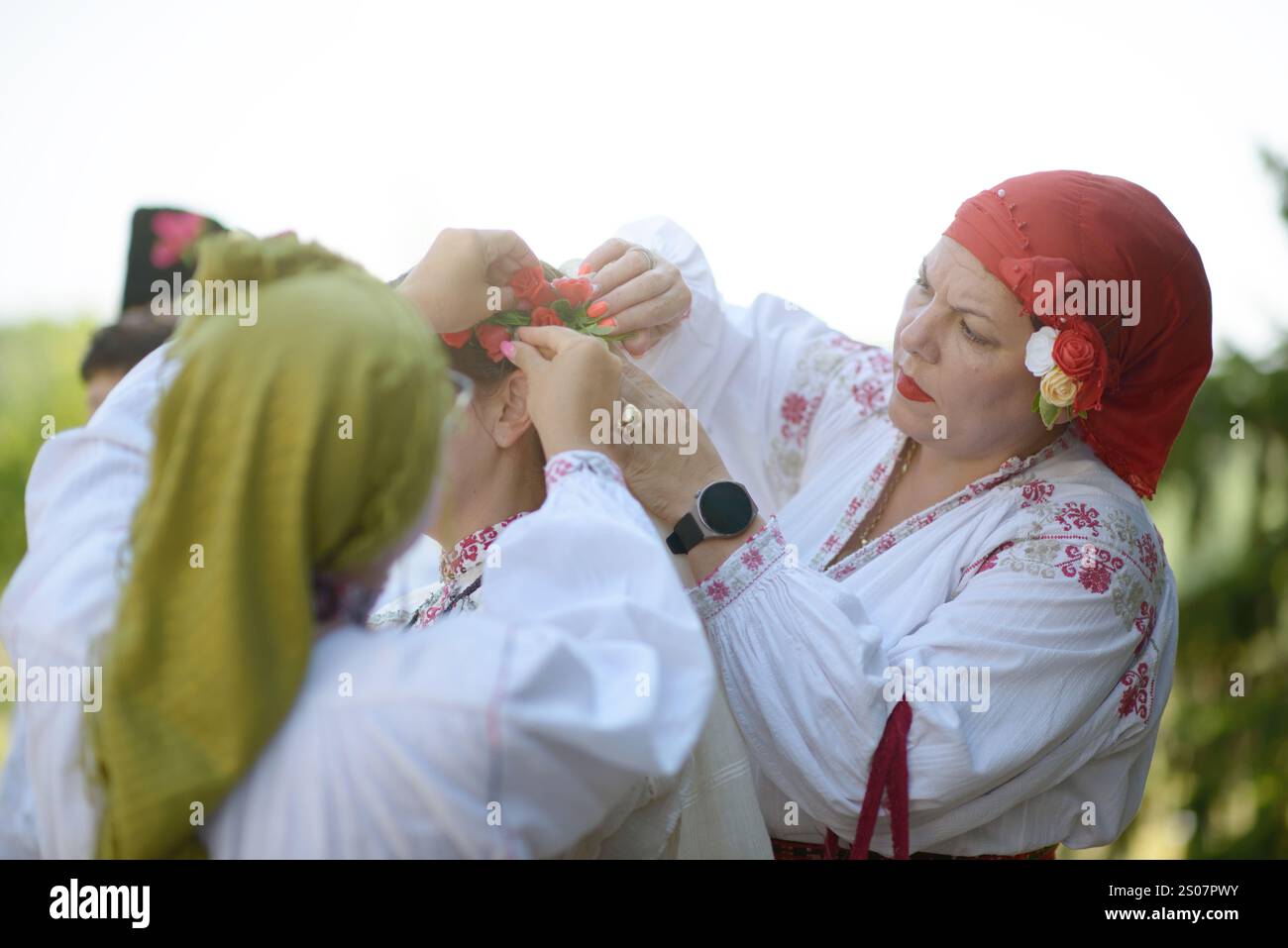 07/27/2024 - Vardim, Svistov, Bulgarie : femme en blouse brodée ajuste la coiffe florale sur une autre. Foulard rouge avec des fleurs montre traditionnel Banque D'Images