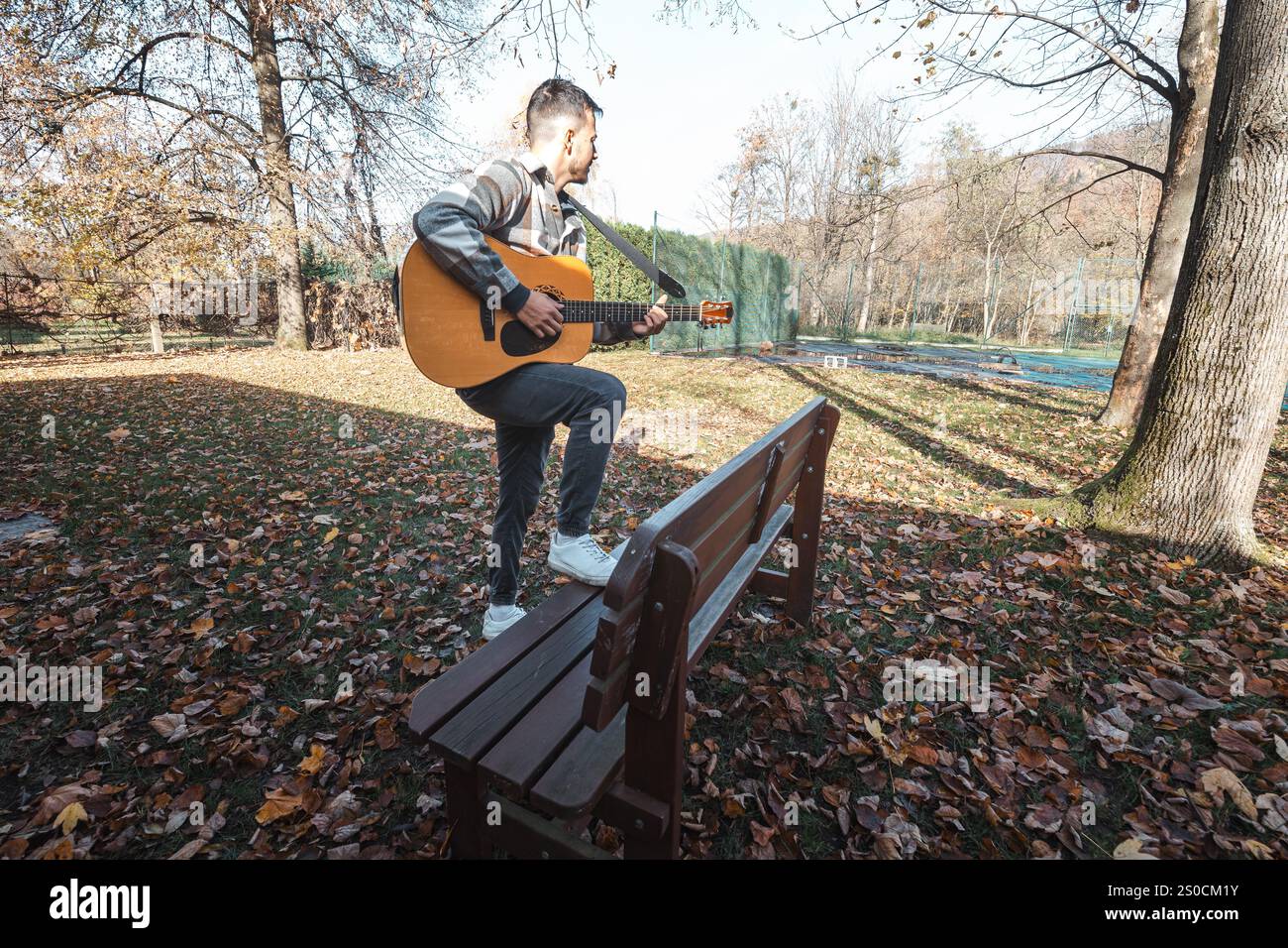 Brun garçon de cheveux assis sur un banc en bois dans un parc d'automne, jouant sur une guitare acoustique, entouré de feuilles tombées colorées, rayonnant un calme et Banque D'Images