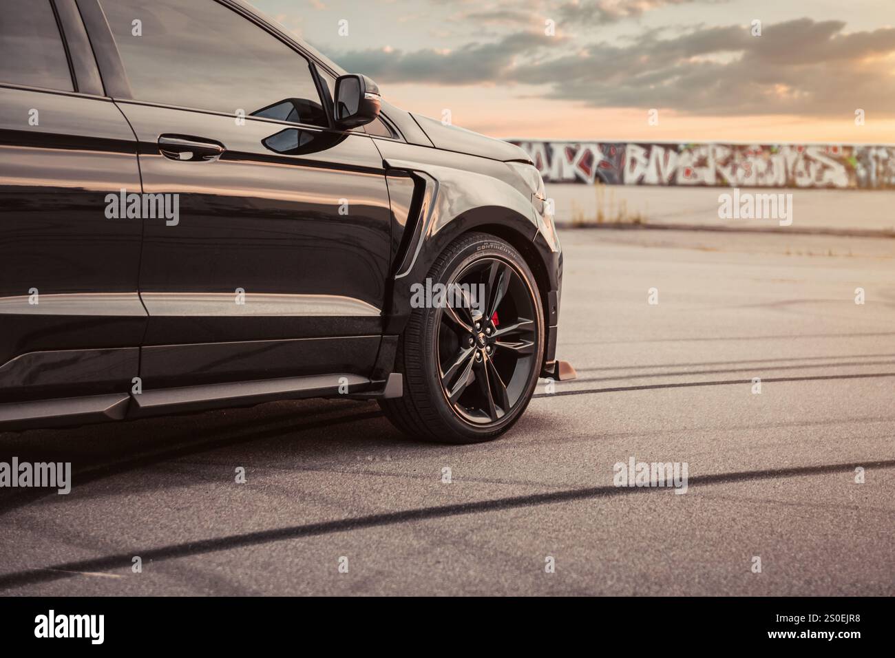 Black Ford Edge ST dans un immense parking au coucher du soleil. Vue de l'aile avant droite d'un VUS sport stationné sur une surface asphaltée avec des marques de pneu, ciel spectaculaire. Banque D'Images