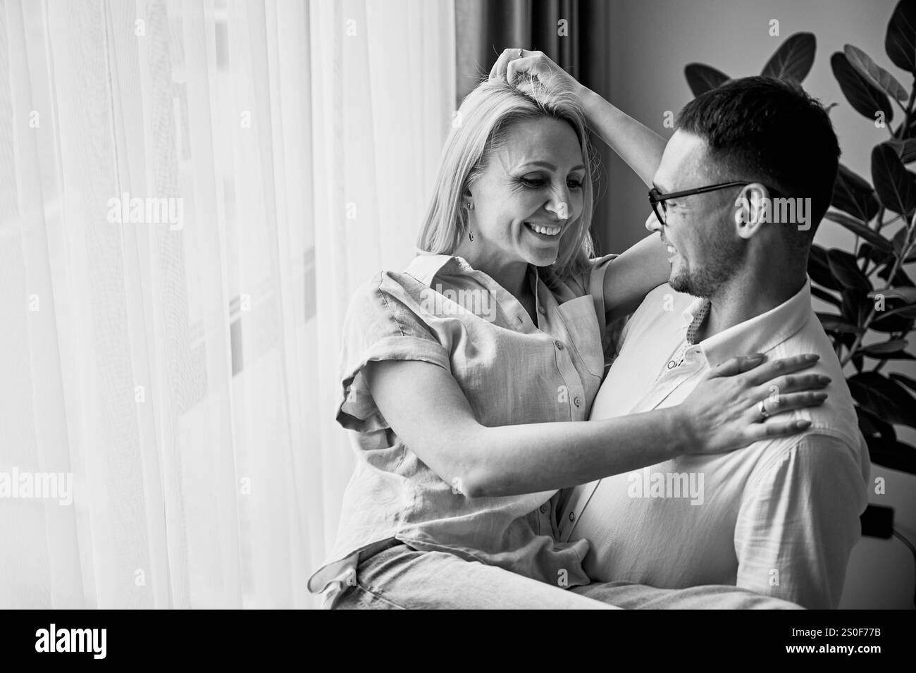 Homme heureux tenant sa femme dans ses bras à la maison. Charmant couple souriant l'un à l'autre. Doux couple se regardant dans des vêtements décontractés sur fond clair. Image en noir et blanc. Banque D'Images