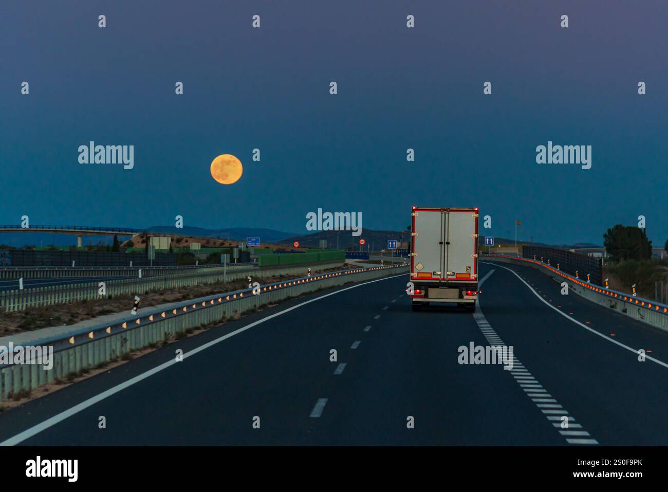 Camion avec une semi-remorque réfrigérée conduisant au crépuscule sur une autoroute sous une pleine lune, vue arrière. Banque D'Images