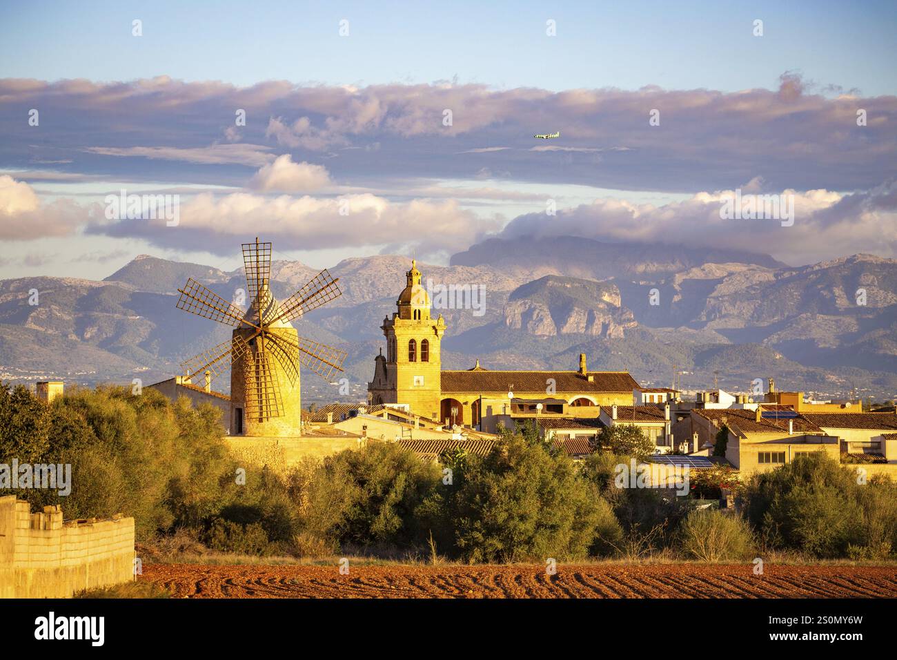 Paysage de montagne avec village, moulin à vent et tour d'église sous un ciel nuageux et avion volant au-dessus des montagnes, Algaida, Majorque, Espagne, Europe Banque D'Images